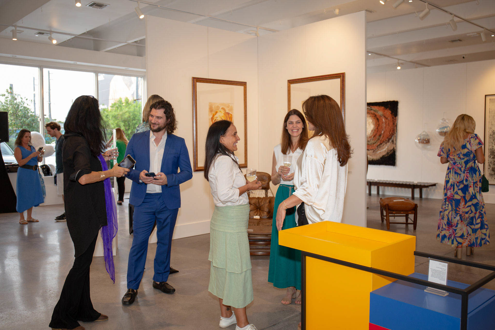 Artist Alex in a blue suit engaged in lively discussion with art collectors at a gallery exhibition, with paintings and sculptures in the background.
