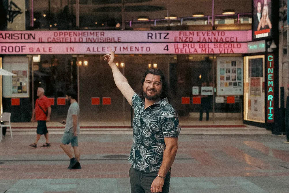 Alex Righetto sorride e indica verso l'alto di fronte all'insegna illuminata del Teatro Ariston al crepuscolo.