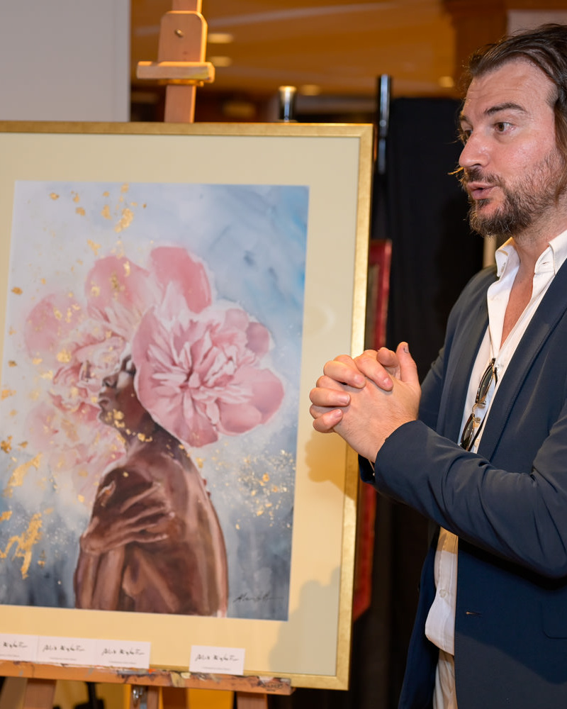 Alex Righetto presenta la collezione Radiance 4 Primo piano di Alex Righetto mentre conversa con un collezionista, con un acquerello della collezione Radiance visibile sullo sfondo durante l’Art & Luxury Monaco 2026.
